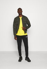 Oliven grøn bomberjakke med to forlommer, gul T-shirt, sorte joggingbukser med logo og sorte sneakers. Simpelt, afslappet outfit.