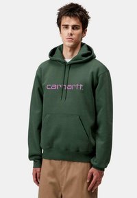 Haut à capuche vert avec une poche kangourou, arborant un logo Carhartt rose. Fabriqué en tissu doux, avec une coupe décontractée et un cordon de serrage ajustable.