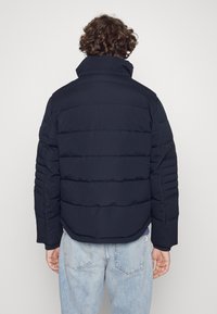 Marinblå puffjacka med kviltad design, hög krage och ribbade detaljer på ärmarna. Bärs med ljusblå denimjeans, vy från baksidan.
