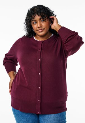 Gilet bordeaux avec un col rond, fermeture à boutons sur le devant, poignets et ourlet côtelés, texture douce, porté sur un haut blanc. Style décontracté.