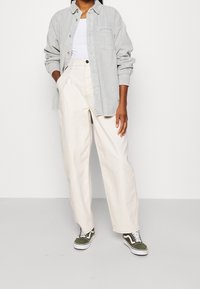 Osoba nosząca jasnoszary sztruksowy koszule, białą koszulkę na ramiączkach, kremowe szerokie spodnie i oliwkowozielone sneakersy z białymi sznurówkami na jednolitym tle.