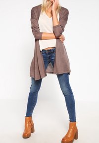 Dlouhý kardigan s otevřeným předním dílem z měkké hnědé látky, kombinovaný s lehkým béžovým topem, tmavě modrými úzkými džíny a hnědými kotníkovými botami na platformě.
