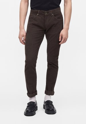 Homme portant un pantalon slim marron retroussé aux chevilles, des chaussettes blanches, des mocassins en cuir noir et une chemise noire sur un fond uni.