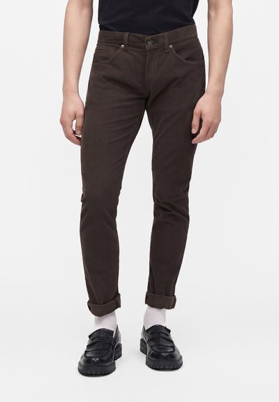 Homme portant un pantalon slim marron retroussé aux chevilles, des chaussettes blanches, des mocassins en cuir noir et une chemise noire sur un fond uni.