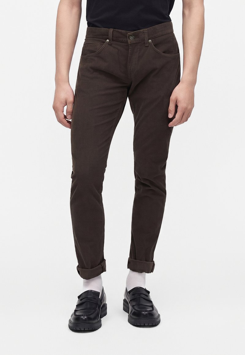 Homme portant un pantalon slim marron retroussé aux chevilles, des chaussettes blanches, des mocassins en cuir noir et une chemise noire sur un fond uni.