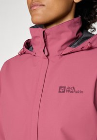 Ljusrosa vattentät jacka med hög krage, med ett svart nätfoder och en logotyp på bröstet. Elastisk dragskor vid huvan.