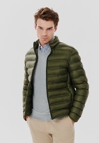 Oliwkowa kurtka puchowa z poziomymi paskami, z zamkiem na przodzie i stojącym kołnierzem. Nosi na sobie jasnoszary sweter i wzorzystą koszulę z guzikami.