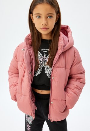 Roze pufferjack met capuchon, voorzien van een ritssluiting en drukknopen. Gedragen over een zwarte trui met een vlindergrafiek en zwarte broek met accenten.