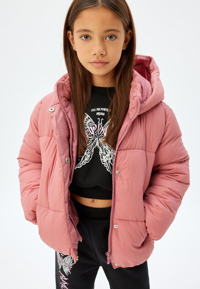 Lyserød dunjakke med hætte, der har lynlås og trykknaplukning. Båret over en sort sweater med en sommerfuglegrafik og sorte bukser med detaljer.