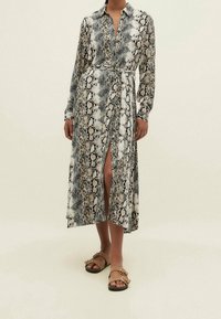 Abito lungo, con bottoni, caratterizzato da una fantasia a serpente nera e beige, con vita cinturata, maniche lunghe e uno spacco laterale. Indossato con sandali marroni.