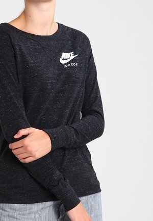 Personne portant un sweat-shirt Nike à manches longues foncé avec le logo "Just Do It", les bras croisés, sur un fond clair uni.