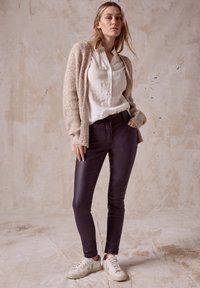 Knitad beige cardigan över en vit skjorta med knappar, parat med mörklila tajta byxor och krämfärgade sneakers. Släta texturer, avslappnad passform.