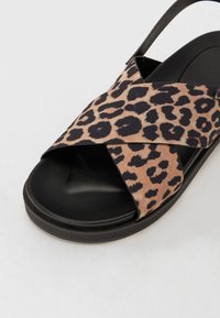 Sandaali leopard-kuosisella ristiinhihnalla, mustalla kumipohjalla ja pehmeällä päällismateriaalilla. Sisältää kevyen kiillon ja teksturoidun pinnan.