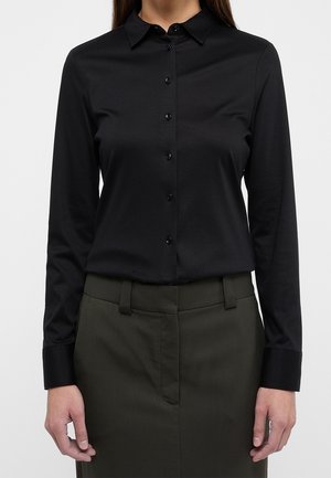 Vrouw met een aansluitend zwart overhemd met lange mouwen en knopen, ingestopt in een hooglopende, donkerolijfgroene broek met ceintuurlussen.