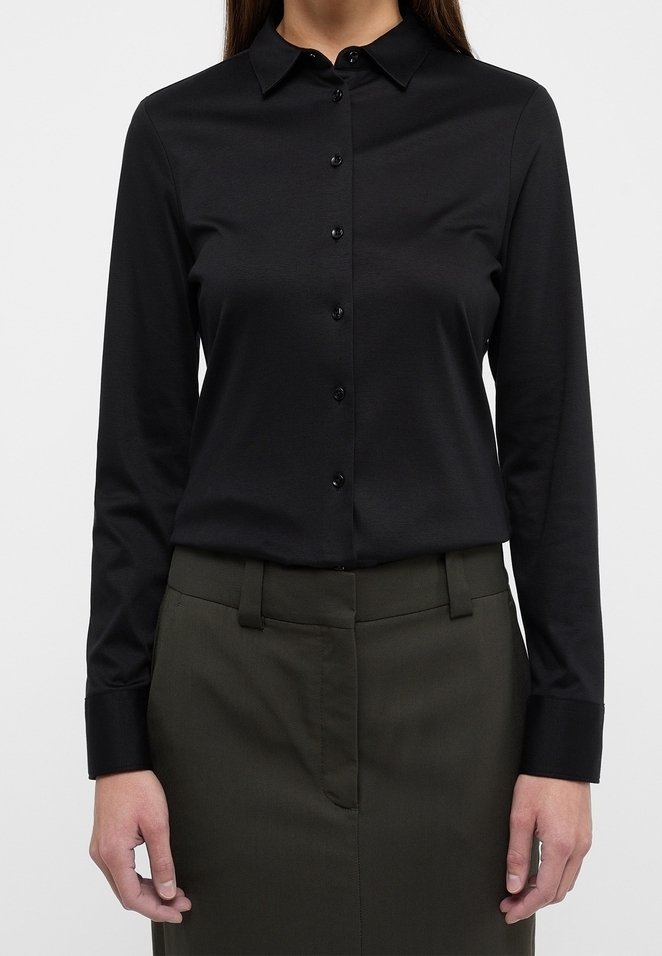 Femme portant une chemise noire cintrée à manches longues avec boutons, rentrée dans un pantalon taille haute olive foncé avec passants pour ceinture.