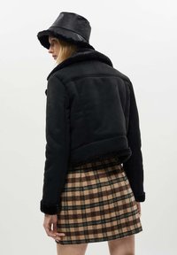 Giacca cropped nera con collare in shearling, abbinata a una gonna a quadri nei toni del marrone e beige. Un cappello a secchiello nero completa l'outfit.