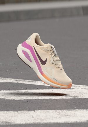 Lyse beige Nike løbesko med lilla og orange detaljer svæver over asfalt med hvide vejmarkeringer.