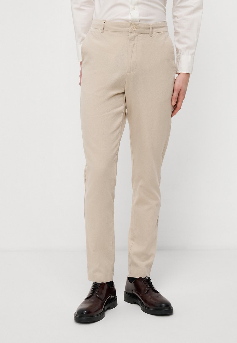 Pantalons beige clair avec une texture lisse, coupe droite et poches avant, associés à des chaussures en cuir brun foncé.