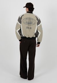 Homme portant une veste bomber beige et grise avec le logo « Prohibited Racing Club Berlin », un pantalon foncé et une casquette marron, debout de dos.
