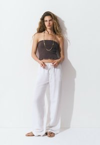 Top corto marrón sin tirantes con detalles fruncidos, pantalones anchos blancos y fluidos, y sandalias beige con tiras finas. El fondo es blanco liso.