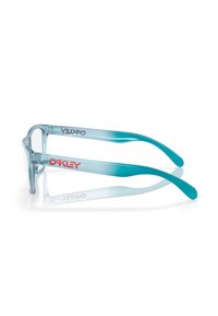 Montatura degli occhiali Oakley trasparente blu con logo Oakley rosso sulle astine, esposta su una superficie riflettente bianca.