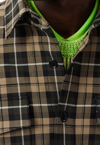 Brun och svart rutigt skjorta med vita linjer, knäppt krage, två framfickor, lager över en ljusgrön stickad tröja.