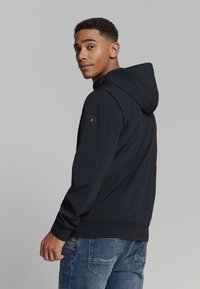 Marineblauwe hooded jack van gladde stof, met een hexagonaal logo-patch op de linker schouder en ribgebreide manchetten. Ggedragen met blauwe jeans.