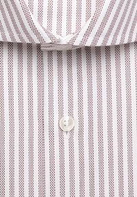 Chemise blanche avec un motif à fines rayures verticales rouges et brunes et un bouton blanc fermé sous le col.