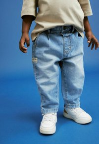 Jeans in denim di colore blu chiaro con una vestibilità rilassata, dotati di un'etichetta a patch sulla tasca posteriore, abbinati a sneaker bianche su uno sfondo blu.