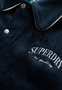 Chemise en velours côtelé marine avec un col à piping blanc et des boutons-pression. Logo "Superdry" brodé en blanc sur le devant.