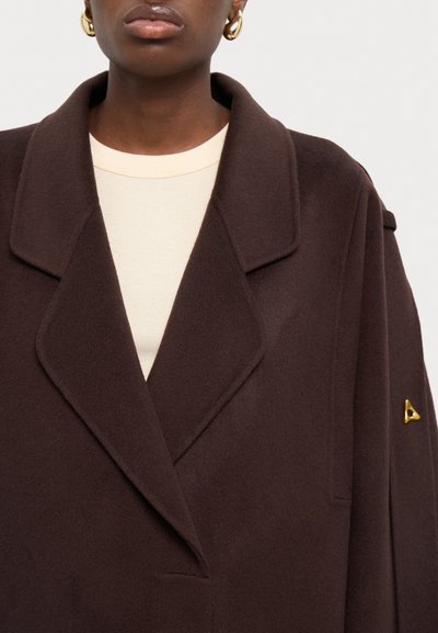 Hnědý kabát ze směsi vlny s výstřihovým límcem, bočními kapsami a zlatým trojúhelníkovým akcentem na rukávu. Textura vypadá jemně a hladce.