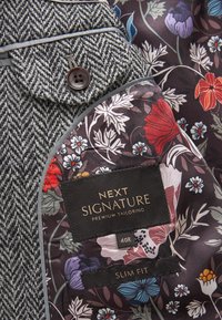 Grijs visgraatstof met contrasterende bloemenvoering. Voorzien van een bruine knoop en labels met maat 40R en slim fit.