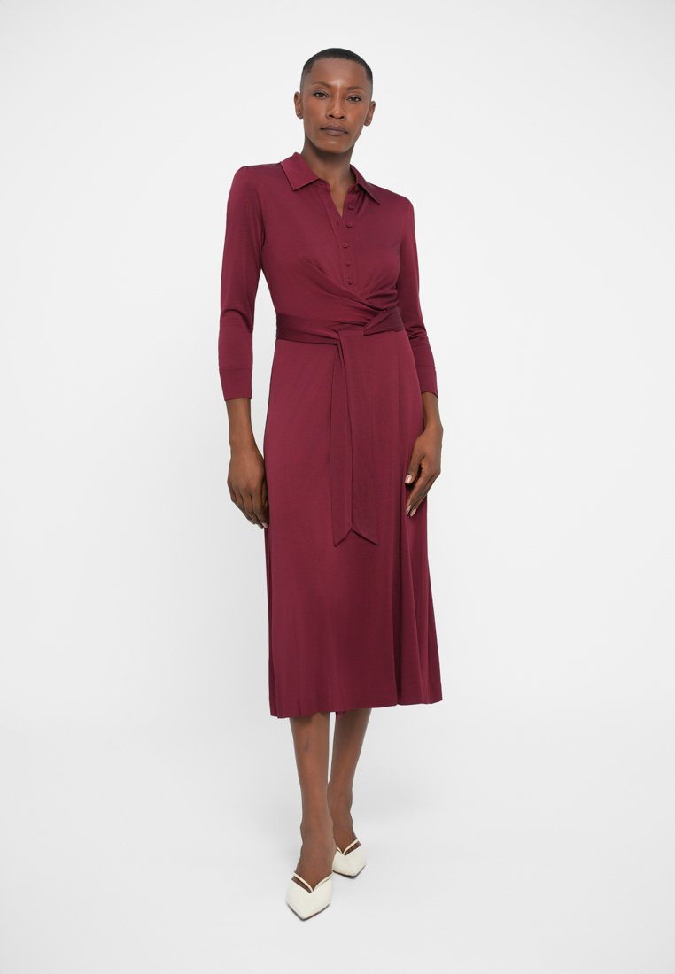 Robe bordeaux avec un col boutonné et des manches trois-quarts. Présente un design enveloppant avec une ceinture à nouer à la taille. Texture lisse.