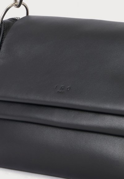 Sac bandoulière en cuir noir avec un anneau métallique argenté et un texte embossé subtil sur le rabat avant.