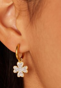Boucle d'oreille en or avec un charme en forme de trèfle à quatre feuilles incrusté de cristaux transparents, mettant en valeur une finition brillante et lisse.