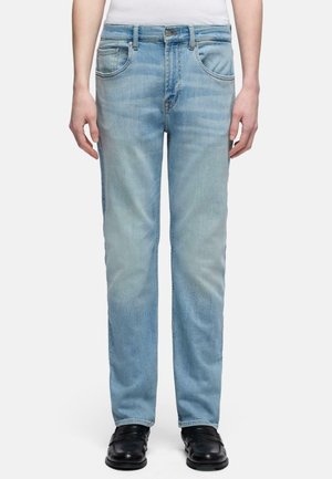 Person trägt hellblaue Jeans mit geradem Bein und Fronttaschen sowie schwarze Schuhe, gezeigt von der Taille bis zu den Füßen vor weißem Hintergrund.