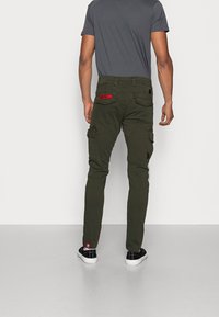Homme portant un pantalon cargo vert foncé avec plusieurs poches et un écusson rouge, assorti d'un t-shirt gris et de baskets basses noires.