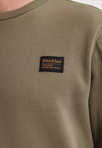 Sudadera verde oliva con una tela texturizada, que presenta una etiqueta tejida en negro y naranja que dice "Petrol Ind." en el lado izquierdo.