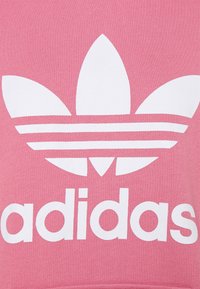 Vitt Adidas trefoil-logotyp och text på rosa tygbakgrund, som visar en del av ett plaggs framsida.