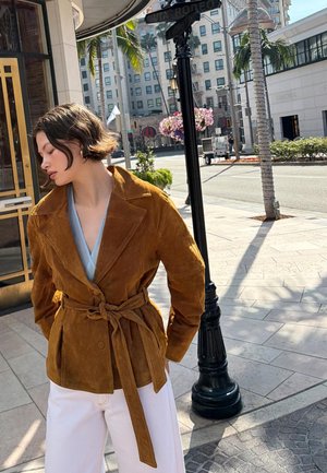 Kobieta w brązowej kurtce z paskiem i białych spodniach stoi na rogu ulicy w mieście obok czarnego słupa z tabliczką i palm.