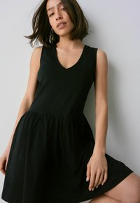 Schwarzes ärmelloses Kleid mit V-Ausschnitt, tailliertem Oberteil und weit schwingendem Rock. Hergestellt aus glattem Material mit einem subtilen Glanz. Keine Muster oder Akzente.