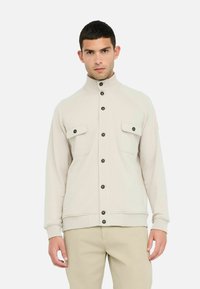Giacca zip-up beige chiaro con colletto alto, bottoni neri e due tasche anteriori a toppa. Tessuto morbido, polsini e orlo a coste.