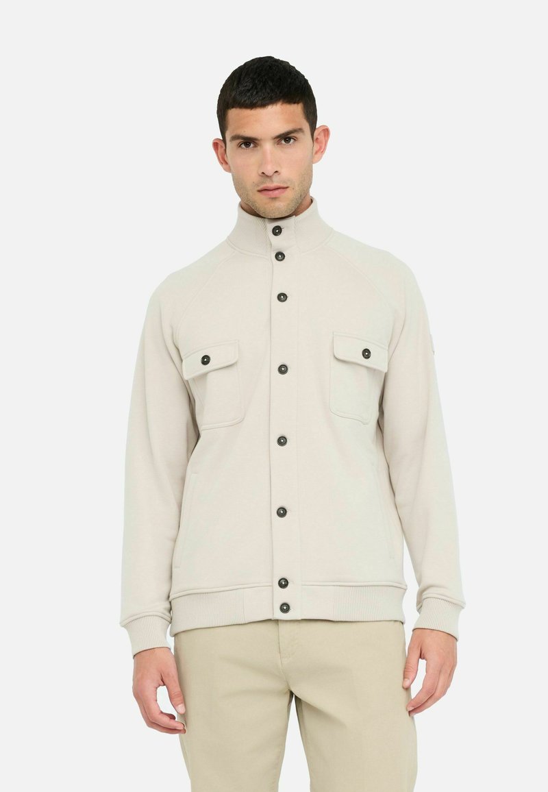 Giacca zip-up beige chiaro con colletto alto, bottoni neri e due tasche anteriori a toppa. Tessuto morbido, polsini e orlo a coste.