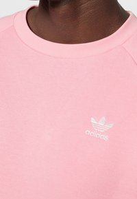 Růžová mikina s kulatým výstřihem, vyrobená z měkké látky. Na hrudi je malé bílé logo Adidas a rukávy typu raglán.