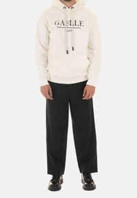 Sudadera crema con el estampado en negro de "GAËLLE" y "PARIS", que presenta un bolsillo delantero y cordones, combinada con pantalones de pierna ancha negros y zapatos.