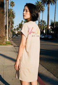 Junge Frau mit kurzen schwarzen Haaren, die ein beiges Kleid mit buntem Text auf dem Rücken trägt und tagsüber auf einem von Palmen gesäumten städtischen Bürgersteig steht.