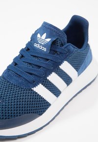 Modrá sportovní obuv s síťovinovým svrškem, semišovými akcenty a bílým trojpruhovým designem. Obsahuje polstrovanou podešev a zavazování na šňůrky.