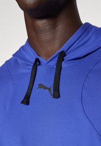 Blauwe hoodie gemaakt van een gladde stof. Heeft een zwart logo en trekkoorden. Close-up opname benadrukt de halslijn en textuur.
