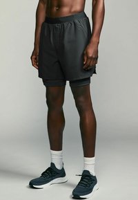 Sorte atletikshorts med elastisk talje, der har et lagdelt design over tætsiddende sorte shorts. Båret med mørke sneakers og hvide sokker.