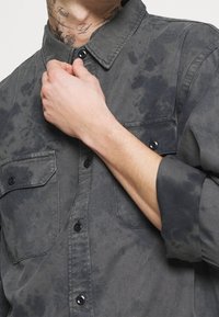 Camicia grigia con bottoni in denim, caratterizzata da un motivo sbiadito e usurato. Presenta due tasche sul petto e bottoni neri, con le maniche arrotolate.